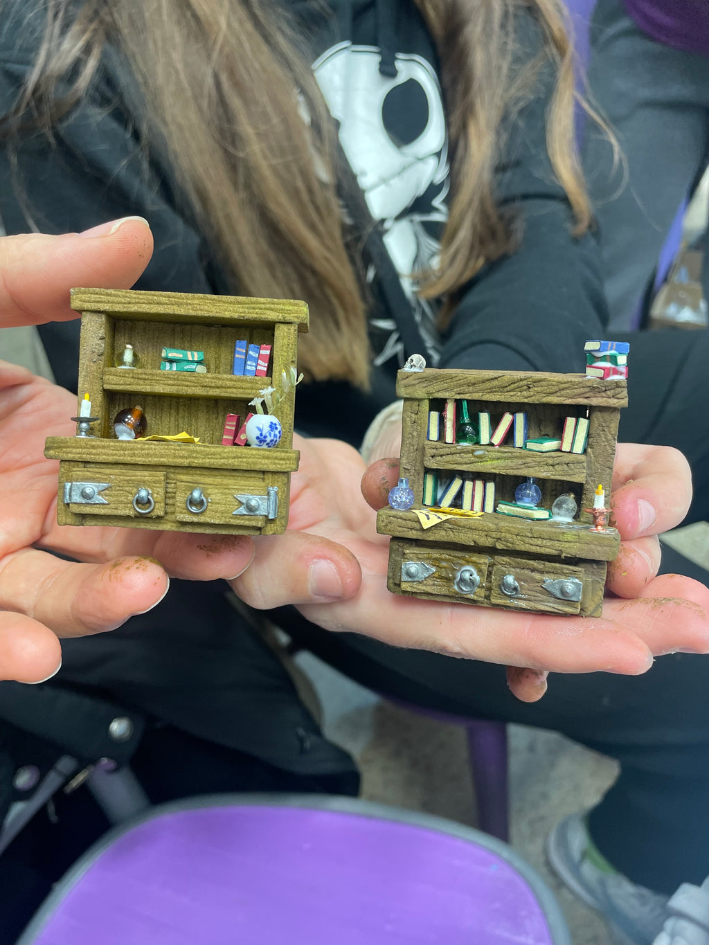 Dos librerías en miniatura sostenidas con las manos, con libros y pequeños accesorios
