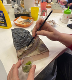 Manos aplicando vegetación con un pincel junto a las rocas de un diorama